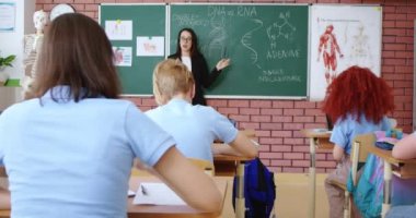 Gözlüklü ve siyah takım elbiseli hevesli bayan okul müdürü biyoloji dersi sırasında öğrencilerin sınıf DNA 'sı ve RNA yapısında rol aldığını anlatıyor. Eğitim ve bilgi kavramı.