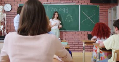 Kahverengi saçlı, ekoloji temalı güzel bir kız. Sınıfta tahtaya yakın duruyor. Erkek öğretmen ve okul arkadaşları başarılı bir konuşma sırasında el çırpıyorlar..