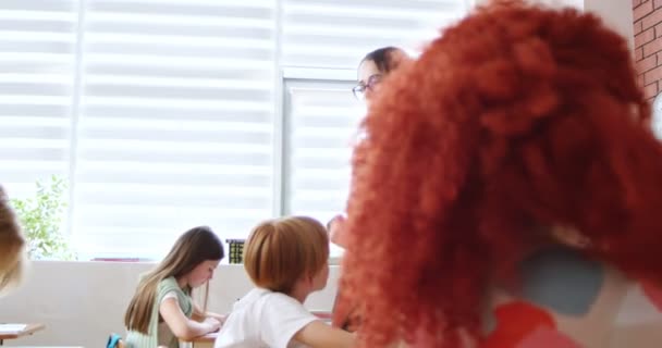白人の厳格な女性教師は 学校でテストに合格する子供たちを見ています 机に座り 検査を書く子供たちの後ろ スタディチェックのコンセプト マスターのコントロール下の男の子 クラスでの試験 — ストック動画