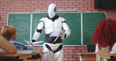 Beyaz modern robot, ilkokul öğrencilerine kontrol işlerini verirken sıraların arasında yürüyor. İnsan emeğinin yapay zekayla değiştirilmesi.