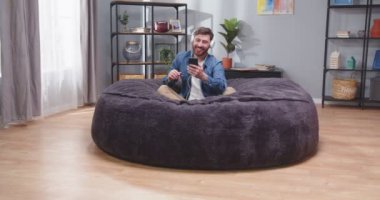 Kanepede elinde akıllı telefonuyla oturmuş kulaklıkla müzik dinleyen yetişkin bir adamın portresi. Kanepede dans eden adam. Erkek, sosyal medyayı zımbırtıda kaydırıyor. Mutluluk kavramı.