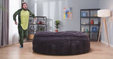 Timsah kostümlü ve sakallı yetişkin bir adam kanepeye zıplıyor. Erkek kameraya bakıp gülümsüyor. Neşeli, mutlu, kaygısız insanlar..