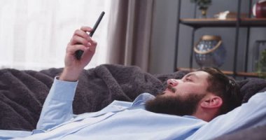 Kanepede uzanmış, kollarını kafasının arkasında kavuşturmuş bir adamın yan portresi. Erkek sosyal medyayı kontrol ediyor. İşten sonra yorgun adam dinleniyor. Dinlenme kavramı, tasasız. Sakallı adamlar..