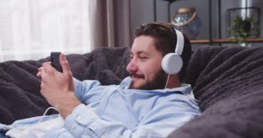 Kanepede elinde akıllı telefonuyla oturmuş kulaklıkla müzik dinleyen yetişkin bir adamın portresi. Adam kanepede dinleniyor. Erkek, sosyal medyayı bir aletle kaydırıyor ve kafasını sallıyor. Dans hareketleri yapıyor..