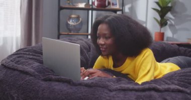 Kanepede dizüstü bilgisayarla yatan Afro-Amerikan kadın portresi. Kadın, aletin üzerinde sosyal medyada geziniyor. İşten sonra kanepede bilgisayar başında dinlenen bir bayan. Yavaş çekim videosu.