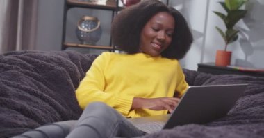 Sarı kazaklı Afro-Amerikan kadın portresi kanepede dizüstü bilgisayarla yazı yazıyor. Kadın dinleniyor ve gülümsüyor. Kadın, aletin üzerinde sosyal medyada geziniyor. İşten sonra kanepede bilgisayar başında dinlenen bir bayan..