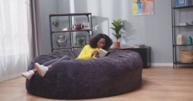 Gözlüklü Afrikalı Amerikalı kadın kanepede uzanıyor, kitap okuyor ve gülümsüyor. Kanepede dinlenen bir kadın. Yalnız zaman geçirmek, kendi kendine çalışmak ve dinlenmek. Karantina konsepti, covid-19.