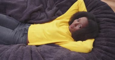 Afro-Amerikan Wooman 'ın kollarını kavuşturup kanepede yatıp etrafa bakınışının en iyi görüntüsü. İşten sonra mutlu bir bayan. Dinlenme kavramı, tasasız. Sarı elbiseli bir kadın..