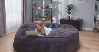 Kanepede dizüstü bilgisayarla yatan yetişkin bir kadının tam bir portresi. Kadın, sosyal medyada bir zımbırtıda kayıyor ve gülüyor. İşten sonra kanepede bilgisayar başında dinlenen bir kadın. Yavaş çekim videosu.