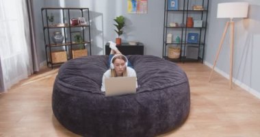 Kadın, kanepeye uzanmış laptopuyla yazı yazıyor kulaklıkla müzik dinliyor. Alet üzerinde sosyal medyada kadın kaydırma ve dans figürleri. Kanepede bilgisayar başında dinlenen bir kadın.