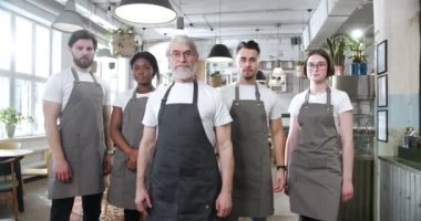 Gri önlüklü çeşitli kafe çalışanları el ele tutuşup gülümsüyor. Geniş açı, kafe çalışanlarının kameraya bakmasına yol açıyor..