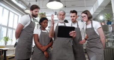 Kafkasyalı eski aşçı elinde tablet cihazıyla aşçıbaşı aşçı ve garsonlardan oluşan ekibiyle yemek siparişleri ve planlama menüsü veriyor. Kafe işi. Restoran işçileri. Önlüklü çok ırklı insanlar..