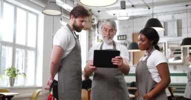 Kafkasyalı yaşlı aşçı elinde tablet cihazıyla genç çoklu etnik garson ve garsonuyla menü hazırlıyor. Kafe işi. Restoran işçileri. Önlüklü ve bilgisayarlı insanlar
