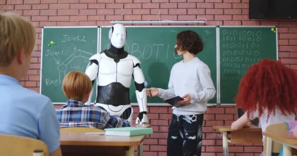 教室の黒板の近くに黒い巻き毛の立っている白人の学校の少年とロボットの操作のためのデジタルタブレットを使用しています 白いヒューマノイドドロイドを与える高い5に弟子にコマンド — ストック動画