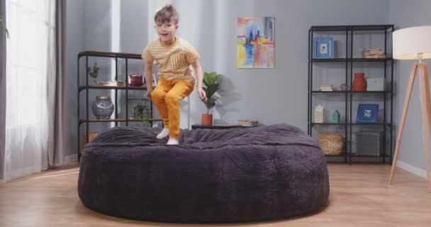 穿着黄色衣服的漂亮男孩在蓬松的沙发上跳着 一个快乐的小男孩被隔离了Coronavirus家的检疫慢动作视频 — 图库视频影像