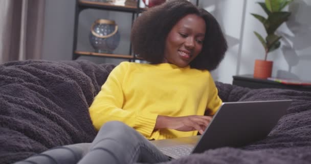 イエローセーターのアフリカ系アメリカ人女性の肖像画は ノートパソコンでタイプするソファーの上に横たわっています 女性が休んで微笑んでいる ガジェットにソーシャルメディアをスクロールする女性 コンピュータでソファーで休憩した後の女性 — ストック動画
