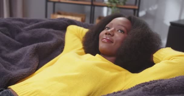 アフリカ系アメリカ人女性が腕でソファーに横たわり 天井を眺めながら頭を横切った 黄色い服を着ている女性 仕事休憩後の疲れた女性 休憩のコンセプト フリー — ストック動画