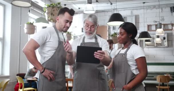 メガネとエプロンのジェスチャーで灰色の髪を被った男は タブレット上の若い労働者の指示を示しています 職場のカフェマネージャーに耳を傾ける多様な従業員 — ストック動画