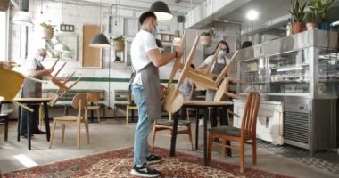 Önlüklü, maskeli, kafeyi temizleyen ve sandalyeleri çıkartan melez insanlar. Çok ırklı erkek ve kadınlar yemek odasını temizliyor. Mataradaki temizleyiciler. Restoran için hazırlık yeri. Yemekten sonra..
