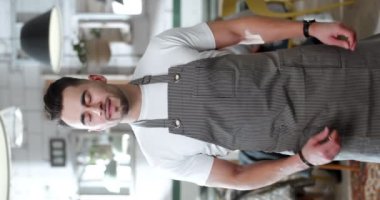 Önlüklü, kafeteryada duran ve kameraya poz veren yakışıklı, beyaz bir genç garsonun dikey portresi. Görüntü formatta. Restoranın erkek şefi..
