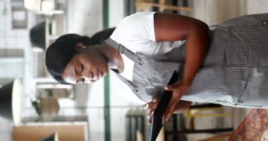 Afro-Amerikalı güzel bayan garsonun dikey portresi. Önlüklü, ayakta duran kafeterya mutfağında. Tablet cihazı kullanıyor ve kameraya gülümsüyor. Restoranın kadın şefi bilgisayar ekranında kayıyor..