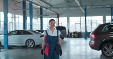 Mavi tulumlu, eldivenli ve koruyucu gözlüklü beyaz bir kadın elinde İngiliz anahtarı ve gülümsemesiyle kameraya doğru yürüyor ve ileriye bakıyor. Geniş bir tamirhanedeki kadın araba tamircisi..