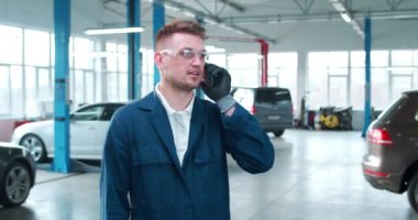 Üniformalı ve gözlüklü yakışıklı beyaz adam oto tamir servisinde cep telefonuyla konuşuyor. Erkek tamirci telefonda sohbet ediyor ve sohbet ediyor. İş günü. Çalışma konsepti.