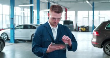 Yakışıklı, beyaz üniformalı, gözlükle takılan, mesaj atan, otomatik tamir servisindeki bilgisayara kayan bir genç. Erkek tamirci tablet kullanıyor. İş günü. Çalışma konsepti.