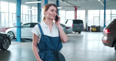 Mavi tulumlu ve beyaz gömlekli genç beyaz kadın geniş bir tamirhanede telefonla konuşuyor, gülümsüyor ve başını sallıyor. İş yerindeki kadın araba tamircisinin portresi.