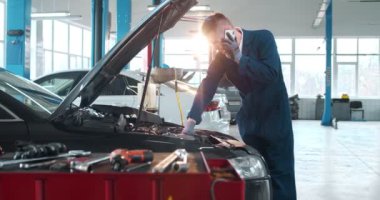 Mavi tulumlu ve siyah eldivenli genç beyaz adam, telefonla konuşurken kaputun altındaki arabayı inceliyor. Erkek tamirci geniş bir tamirhanede çalışıyor..