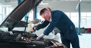 Mavi tulumlu, eldivenli ve koruyucu gözlüklü genç beyaz adam el feneriyle kaputun altındaki arabayı inceliyor. Erkek araba tamircisi geniş bir tamircide çalışıyor..