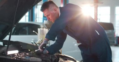 Üniformalı beyaz erkek tamirci araba hurdacısıyla araba tamiri yapıyor. Genç adam motoru tamir ediyor ve büyük garajda anahtarı çeviriyor. Konsepti korumak için. Tamir işi..