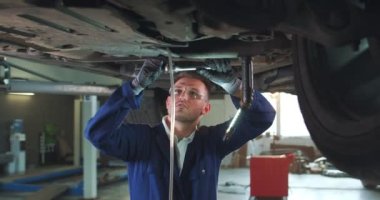 Mavi tulumlu, eldivenli ve koruyucu gözlüklü genç beyaz adam anahtarı kullanarak arabanın altına sürgüyü sıkıyor. Geniş garajda çalışan erkek araba tamircisi..