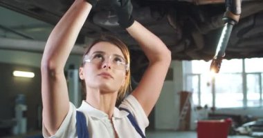 Beyaz kadın tamirci, araba yıkama servisindeki hurdayla arabanın altında tamir yapıyor. Genç kadın mühendis motoru tamir ediyor ve yeraltından İngiliz anahtarını çeviriyor. Konsepti korumak için. Tamir işi..