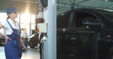 Otomobil servisinde çalışan beyaz kadın tamirci. Büyük garajda yükselen genç bir kadın mühendis. Konsepti korumak için. Tamir için yükseltiyorum. Otomobil kaldırılıyor.