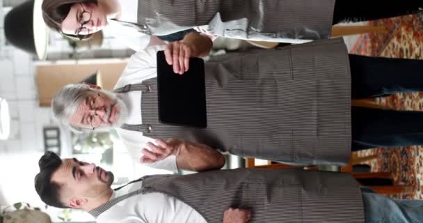 戴眼镜 头戴围裙 留着白胡子的男人给年轻员工展示平板电脑的说明书 男性和女性员工在工作场所聆听咖啡店经理谈话的垂直镜头 — 图库视频影像
