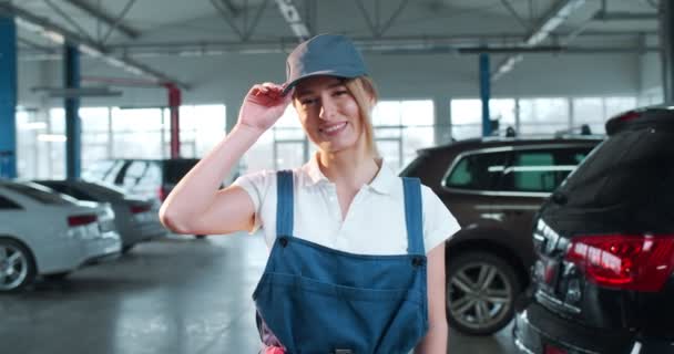 Young Caucasian woman in blue overalls and cap looks into camera, while smiling and giving greeting gesture. Female car mechanic at workplace in spacious repair shop.