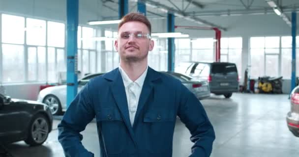 青いカバーオールと保護眼鏡の若い白人男性は 手を渡り 笑顔を浮かべ カメラを見ています 広々とした修理工場の職場での男性車のメカニック — ストック動画