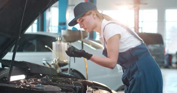 青いオーバーオールの若い白人女性は オープンフードで車に向かって歩き 懐中電灯でエンジンを検査します 広々とした修理工場で働く女性車メカニック — ストック動画