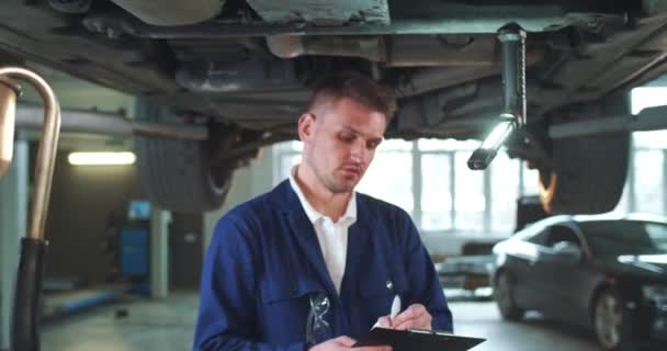 Jonge Blanke Man Blauwe Overall Werkt Onder Auto Ruime Reparatiewerkplaats — Stockvideo