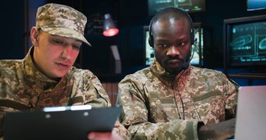 Melez ırk erkek işçileri izleme merkezinde oturup çatışma bölgelerinden gelen verileri inceliyor. Kafkasyalı asker belge okurken, kulaklıklı Afrikalı Amerikalı bir adam dizüstü bilgisayarda yazı yazıyor.