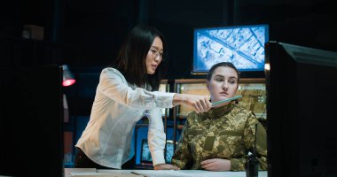 Askeri kontrol ofisinde okuyan asker üniformalı beyaz bir genç kız. Asyalı bayan öğretmen orduda kadınlara öğretmenlik yapıyor ve izleme odasında bilgisayar ekranında bir şey gösteriyor..