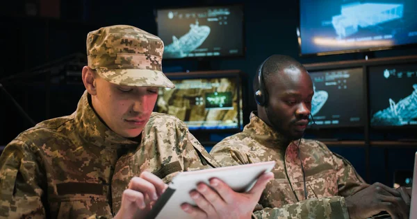African American young man in headset typing on laptop computer and Caucasian soldier tapping on tablet device in control room. Multiethnic army officers in uniforms working together in office.