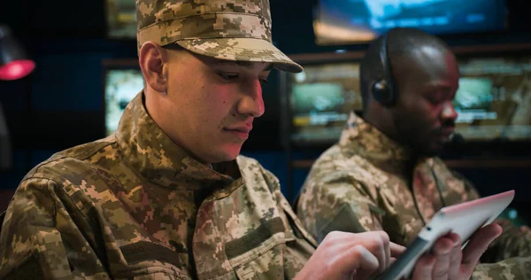 Close up of Caucasian man soldier in uniform and cap tapping, scrolling and surfing online on tablet device in monitoring room. African American male colleague in headset working on background.