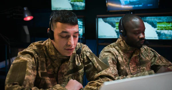 Mixed-races male dispatchers in army co-workers in headsets sitting in military technical room with screens and controlling war operation. Two mixed-races men cooperating in troops control room.