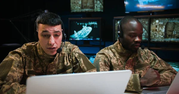 Multiethnic military men in uniforms and headsets talking and videochatting while working as dispatchers. Soldiers sitting at computers and speaking in controlling room.