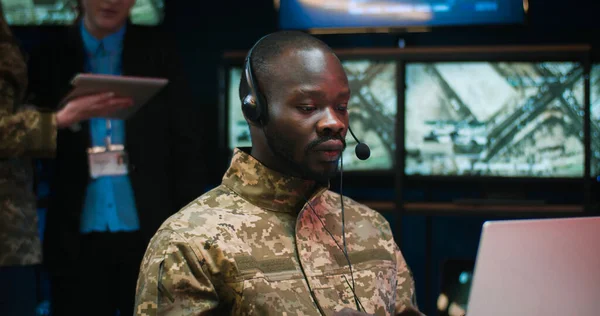 African American military man in headset sitting at laptop computer and talking via videochat in controlling center of army. Soldier speaking via webcam in military office. Officer in dispatch room.