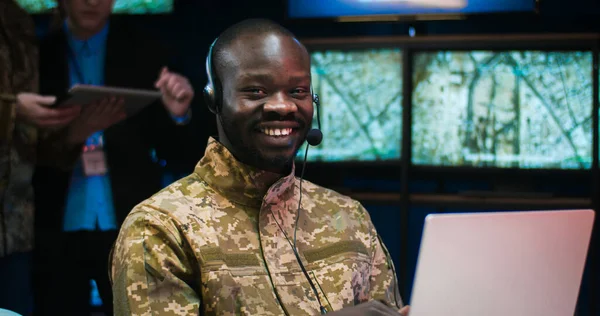 Portrait of African American military man in headset sitting at laptop computer in controlling center of army and smiling joyfully to camera. Male soldier in military office. Officer in dispatch room.