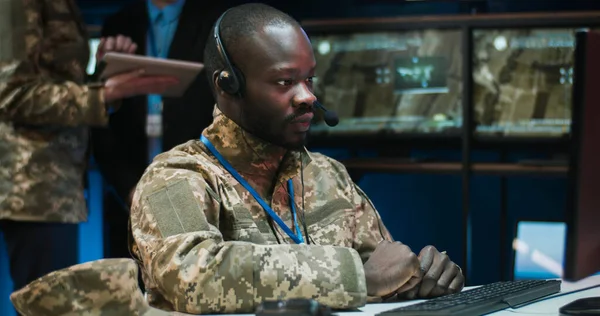 Young African American male military officer in uniform and headset with mic sitting in front of computer and talking via web cam in military call center. Man in army speaking on video call.