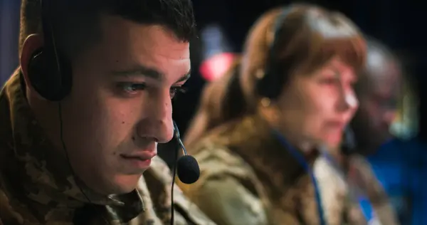 Close up of Caucasian male soldier in headset working as dispatcher in conflict zone controlling office. Blurred woman and African Ameican man colleagues on background.